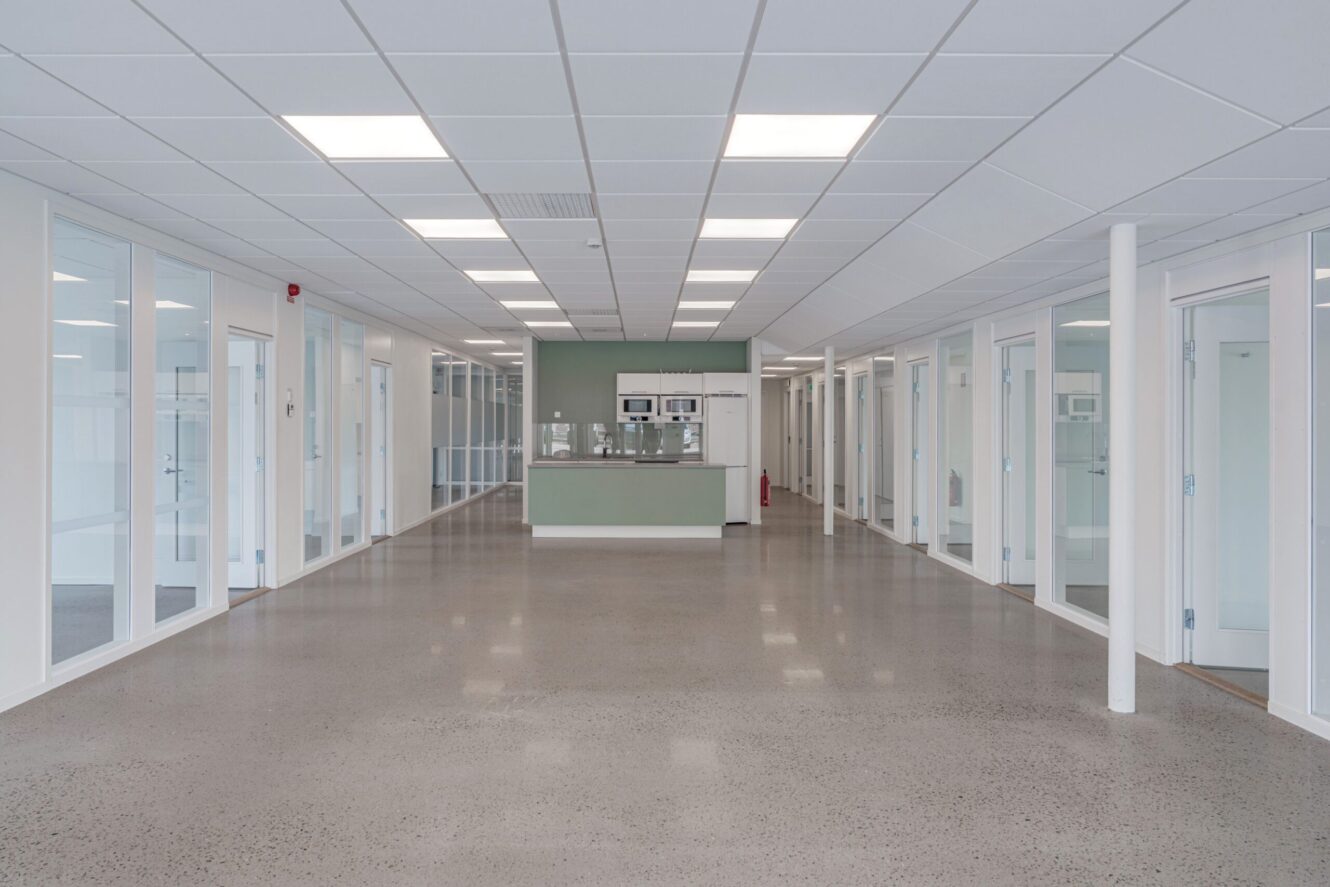 En modern kontorshall med polerade betonggolv, kontor med glasväggar på båda sidor och ett pentry i slutet.