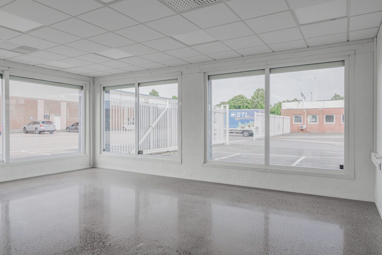 Tomt, omöblerat rum med stora fönster, vita väggar och ett polerat betonggolv, med utsikt över en parkeringsplats och närliggande kommersiella byggnader.