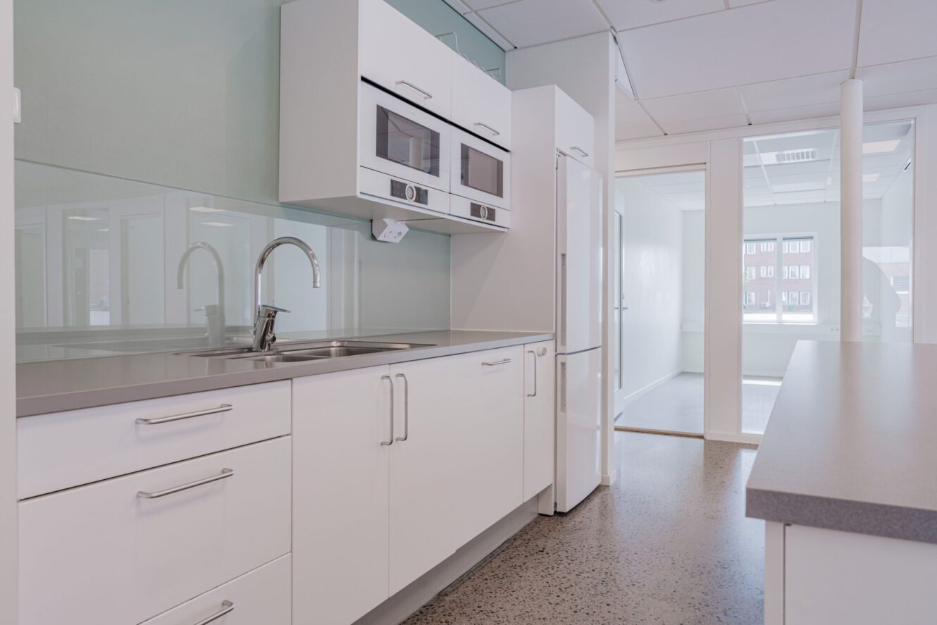 Modernt kök med vita skåp, inbyggd mikrovågsugn, diskho i rostfritt stål, kylskåp och stora fönster som släpper in naturligt ljus.