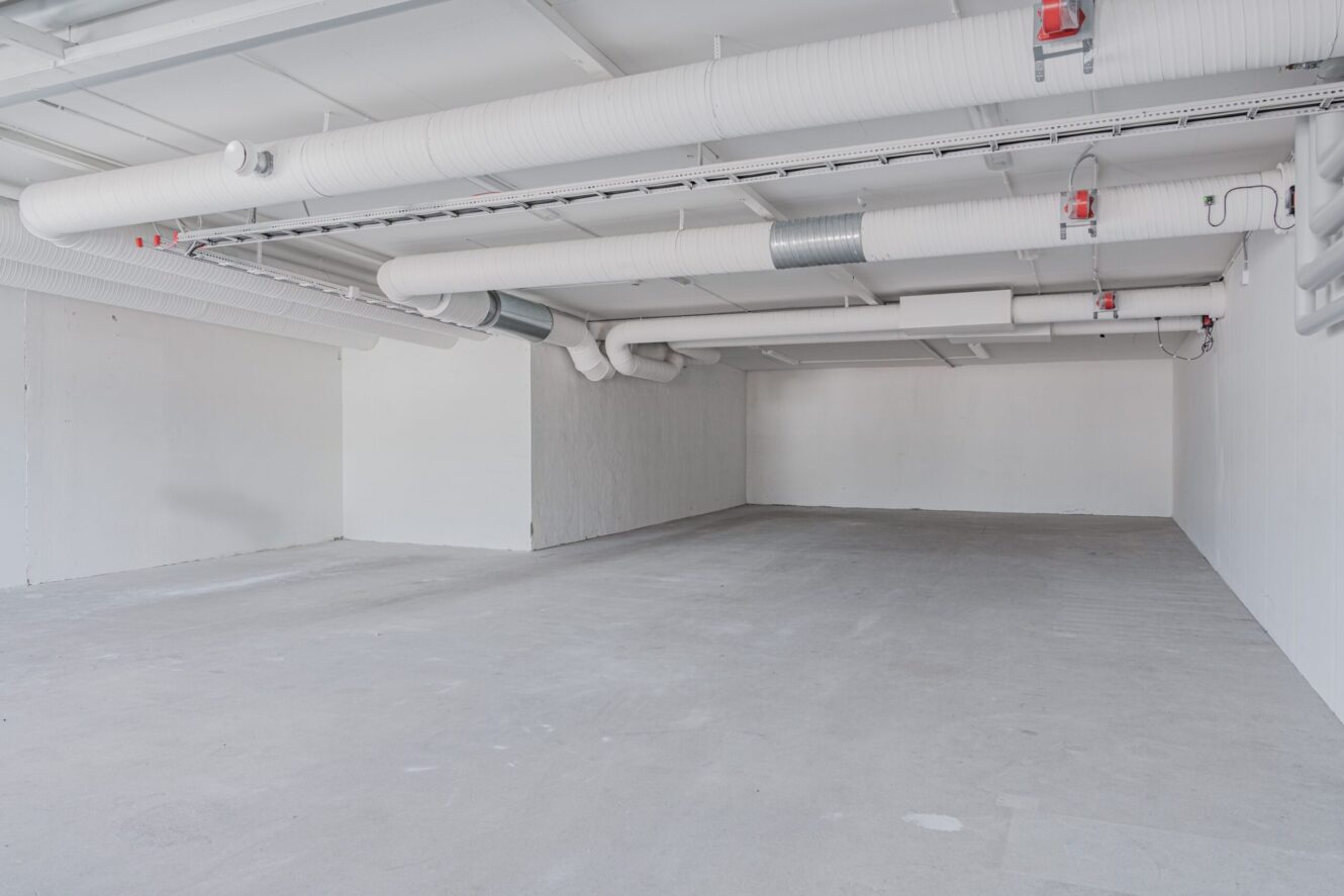 Tomt industriområde med vita väggar, synliga rör i taket och betonggolv, jämnt upplyst av taklampor.
