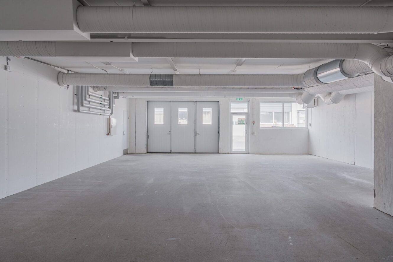 Tomt industrirum med vita väggar, synliga rör i taket, grått golv och tre dörrar med fönster på baksidan som släpper in dagsljus.