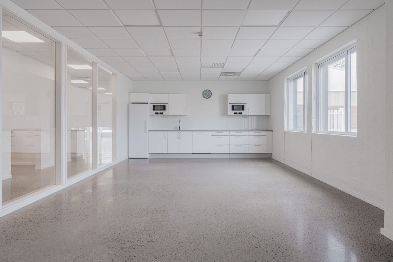 Ett rent, tomt kök med vita skåp, vitvaror och bänkskivor, stora fönster till höger och skiljeväggar av glas till vänster.