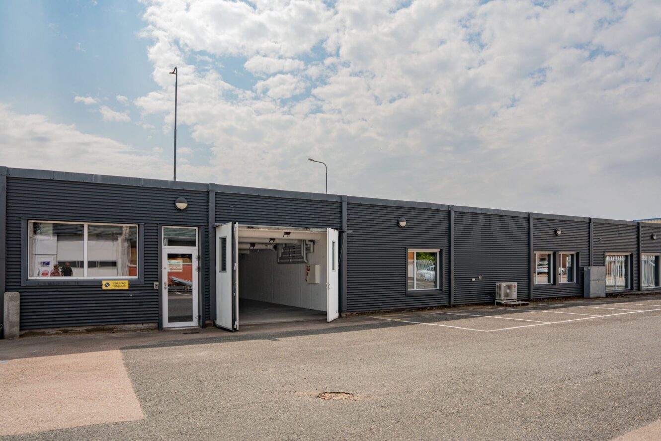 En envånings industribyggnad med grå metallfasad, flera fönster, en öppen garageport och en tom parkeringsplats framför under en delvis molnig himmel.