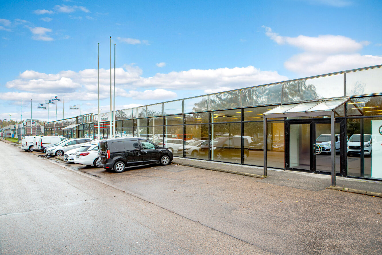 En rad parkerade bilar utanför en lång affärsbyggnad med glasfasad en molnig dag.