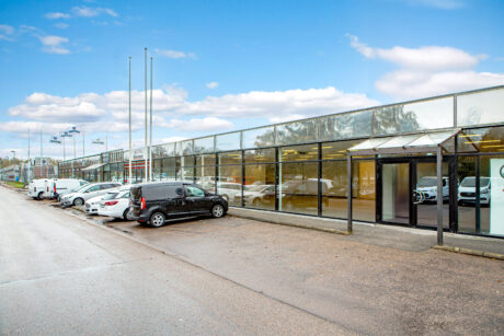En rad parkerade bilar utanför en lång affärsbyggnad med glasfasad en molnig dag.