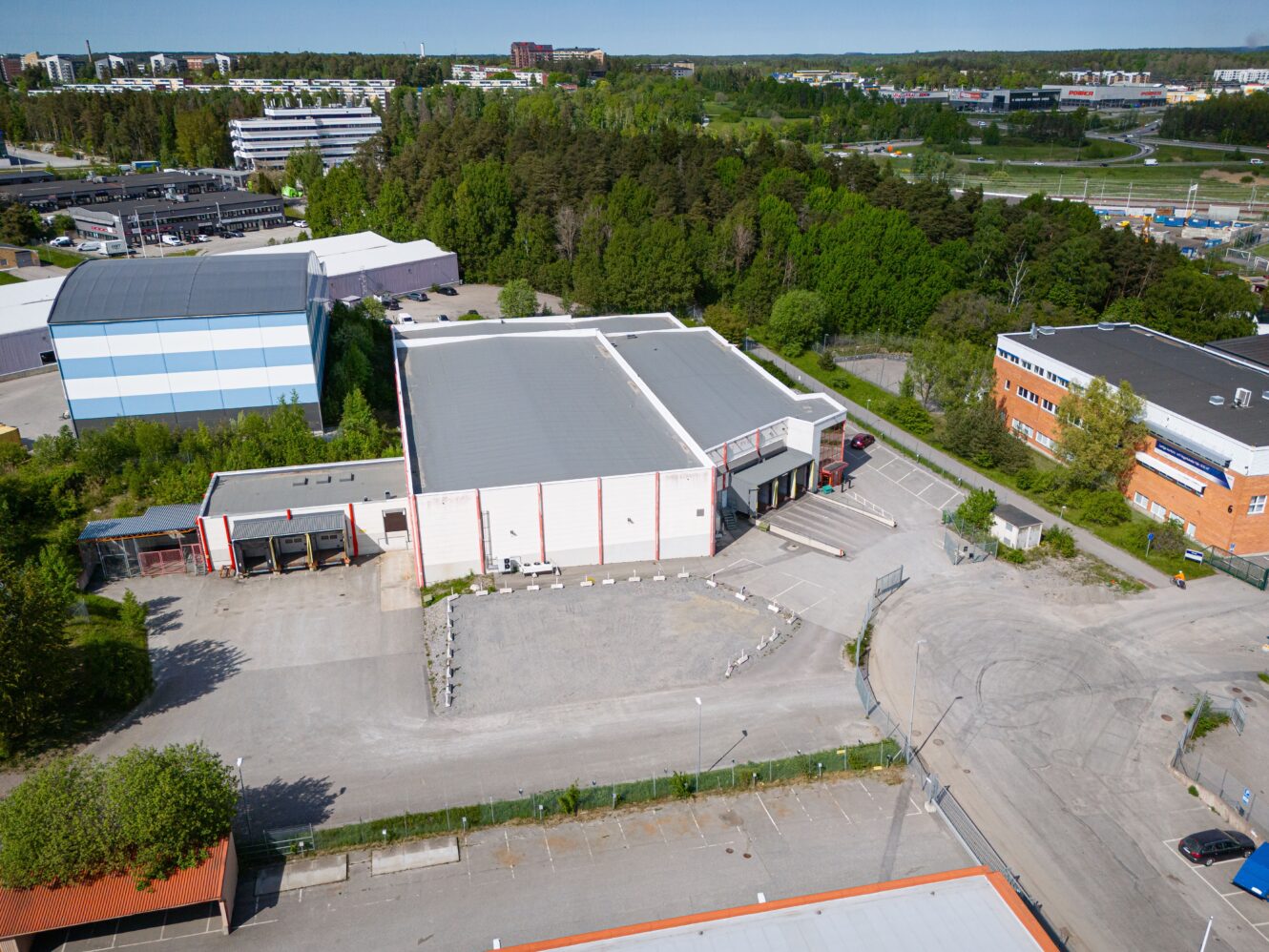 Flygfoto över ett industrikomplex med stora lagerlokaler, parkeringsplatser och omgivande träd, nära vägar och andra byggnader.