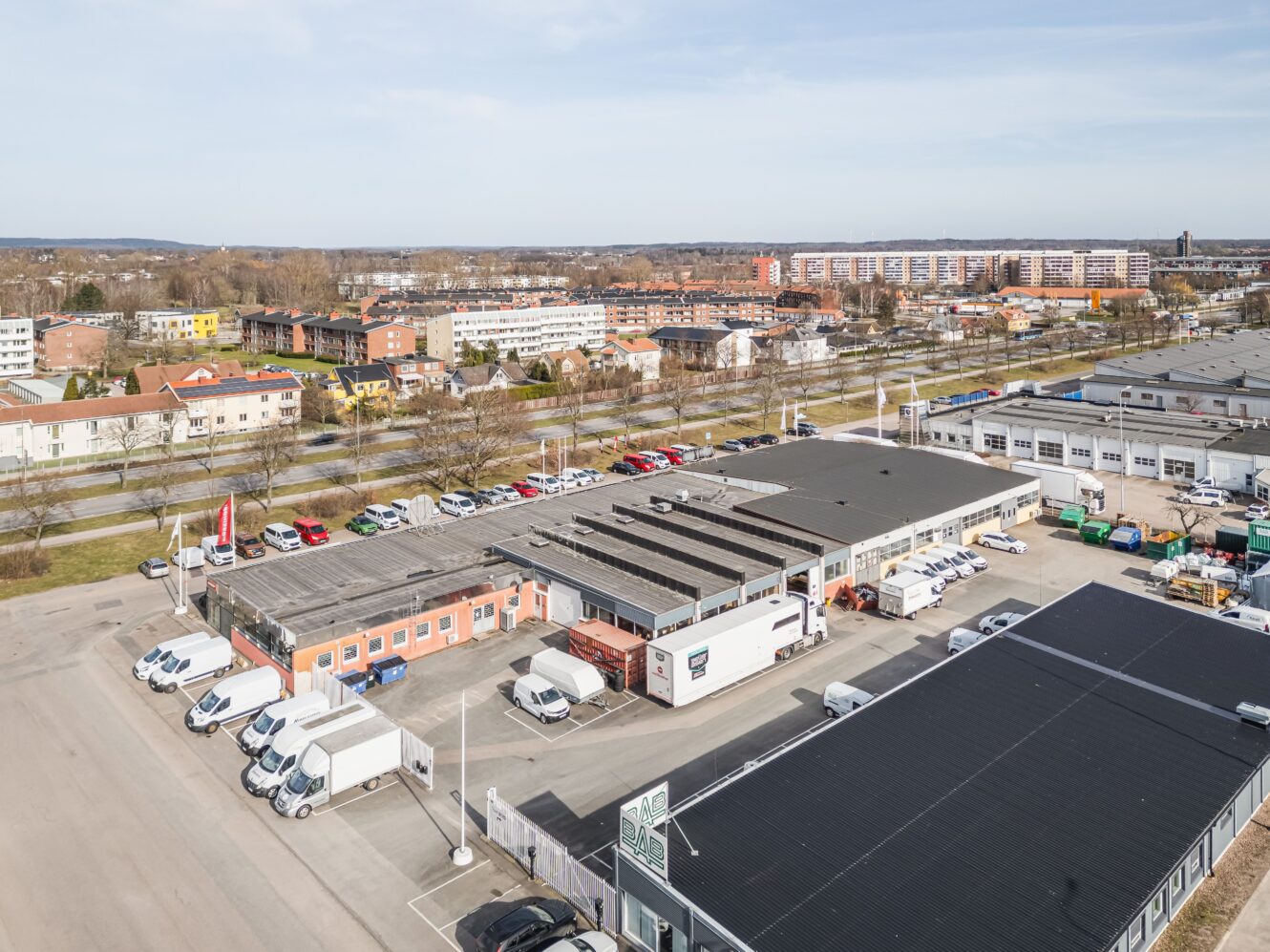 Flygfoto över ett affärsområde med flera byggnader, parkerade vita skåpbilar och bilar samt ett bostadsområde i bakgrunden.