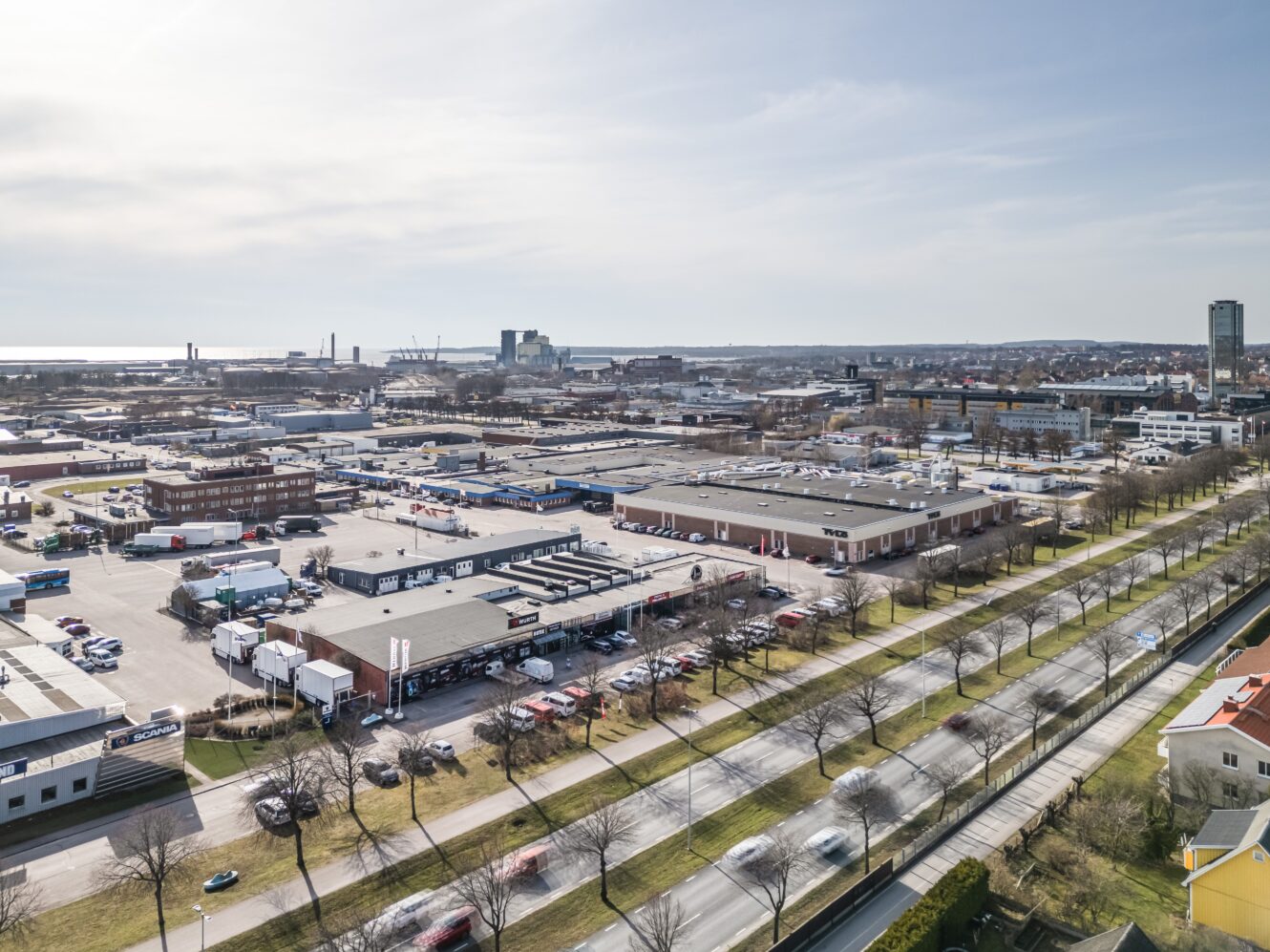 Flygfoto över ett industriområde med lagerlokaler, kontorsbyggnader, parkerade fordon och en flerfilig väg med bilar och träd, med en stadsbild i bakgrunden.