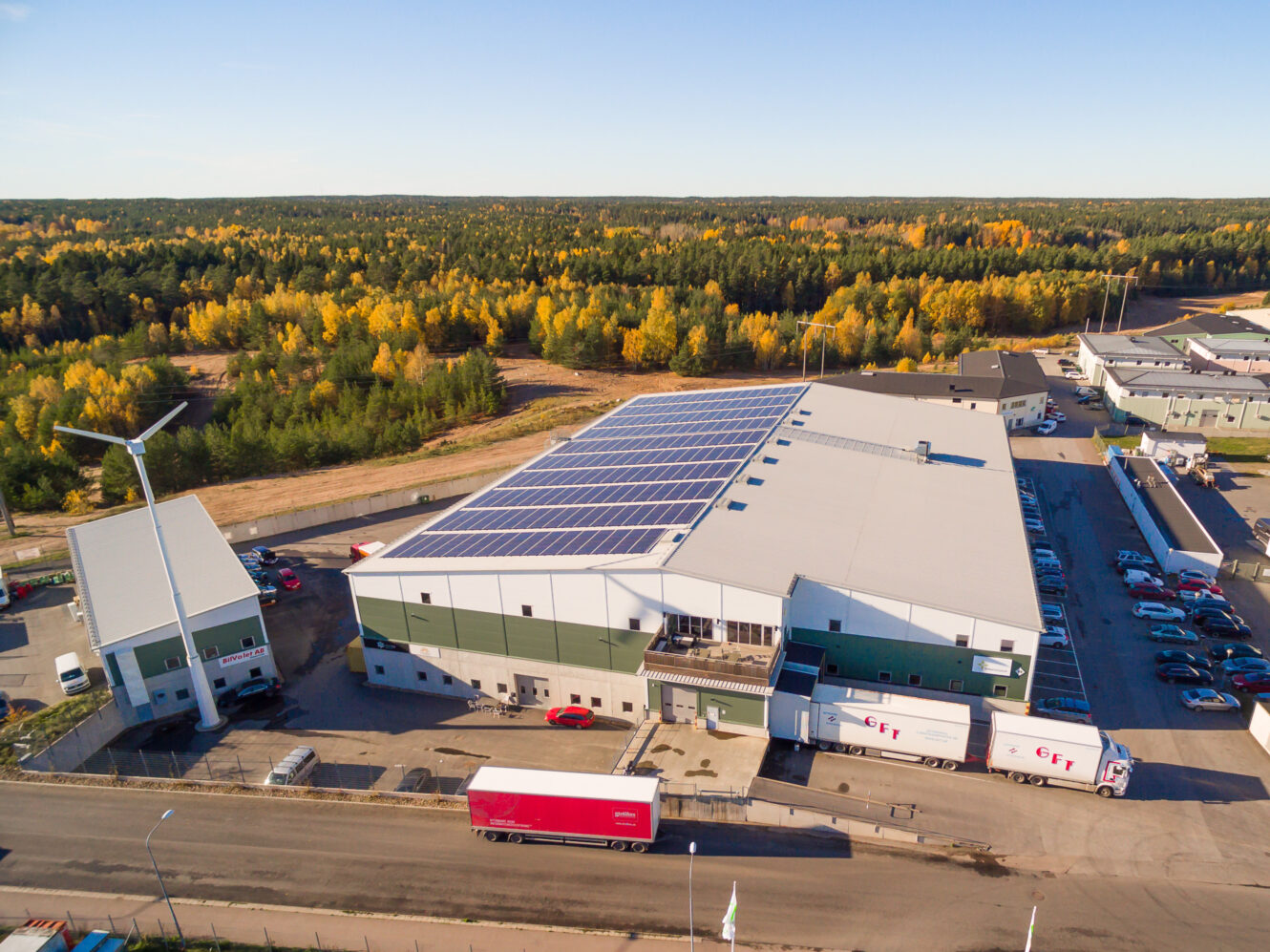 Flygfoto över en stor lagerlokal med solpaneler på taket, omgiven av lastbilar, bilar och ett vindkraftverk, med en skog i bakgrunden.