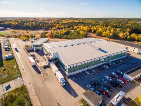 Flygfoto över en stor lagerlokal med lastkajer, lastbilar och parkerade bilar omgivna av träd med höstlöv.