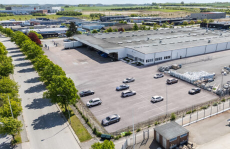 Flygfoto över en stor industrihall med en i stort sett tom parkeringsplats, flera parkerade bilar och omgivande träd och vägar.