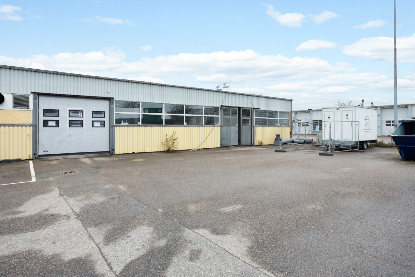 Industribyggnad i en våning med flera garageportar, stora fönster och en asfalterad tomt framför. En fraktcontainer och en sopcontainer syns till höger.