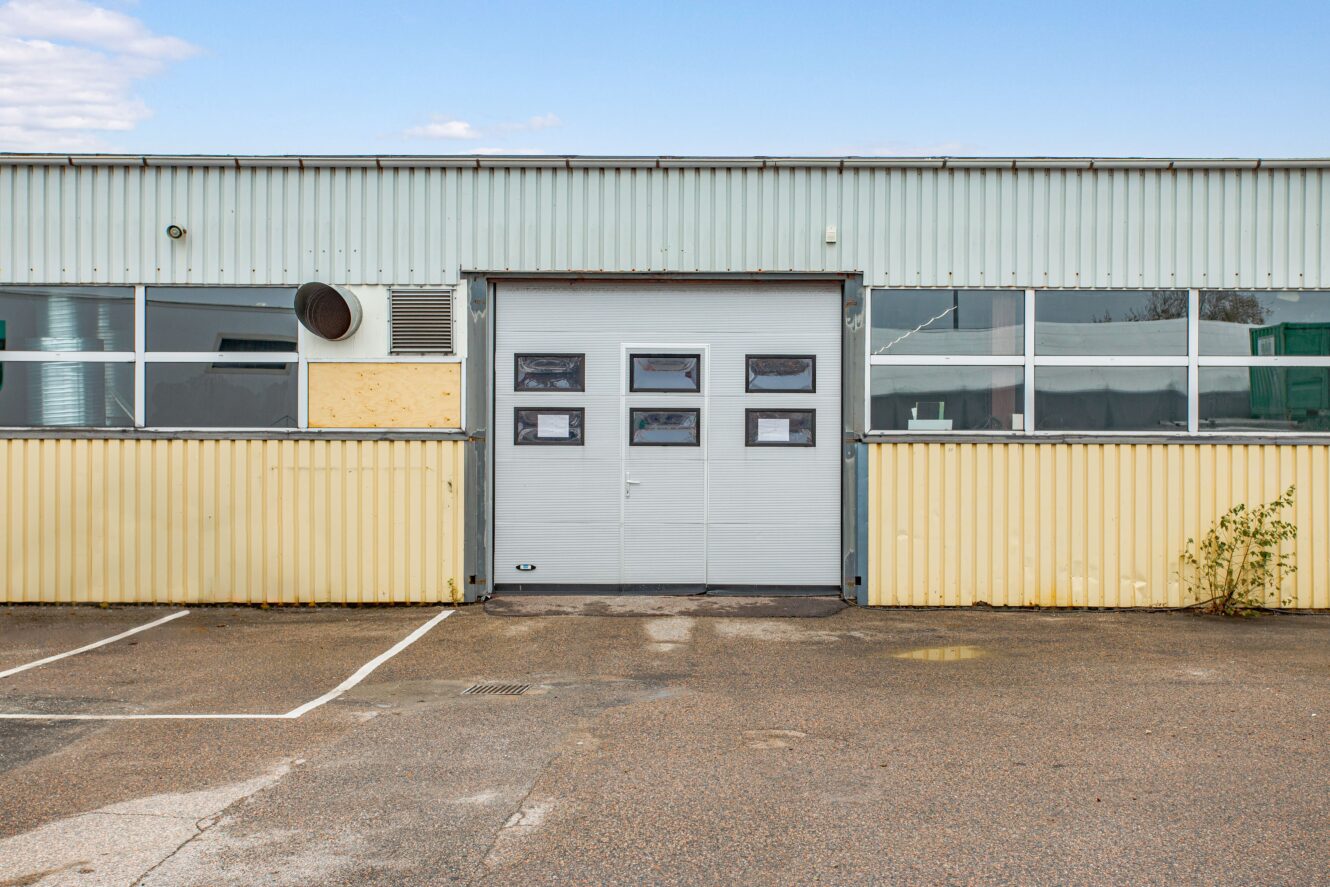 En industribyggnad i korrugerad metall med en central garageport, små fönster och en asfalterad parkeringsplats framför.