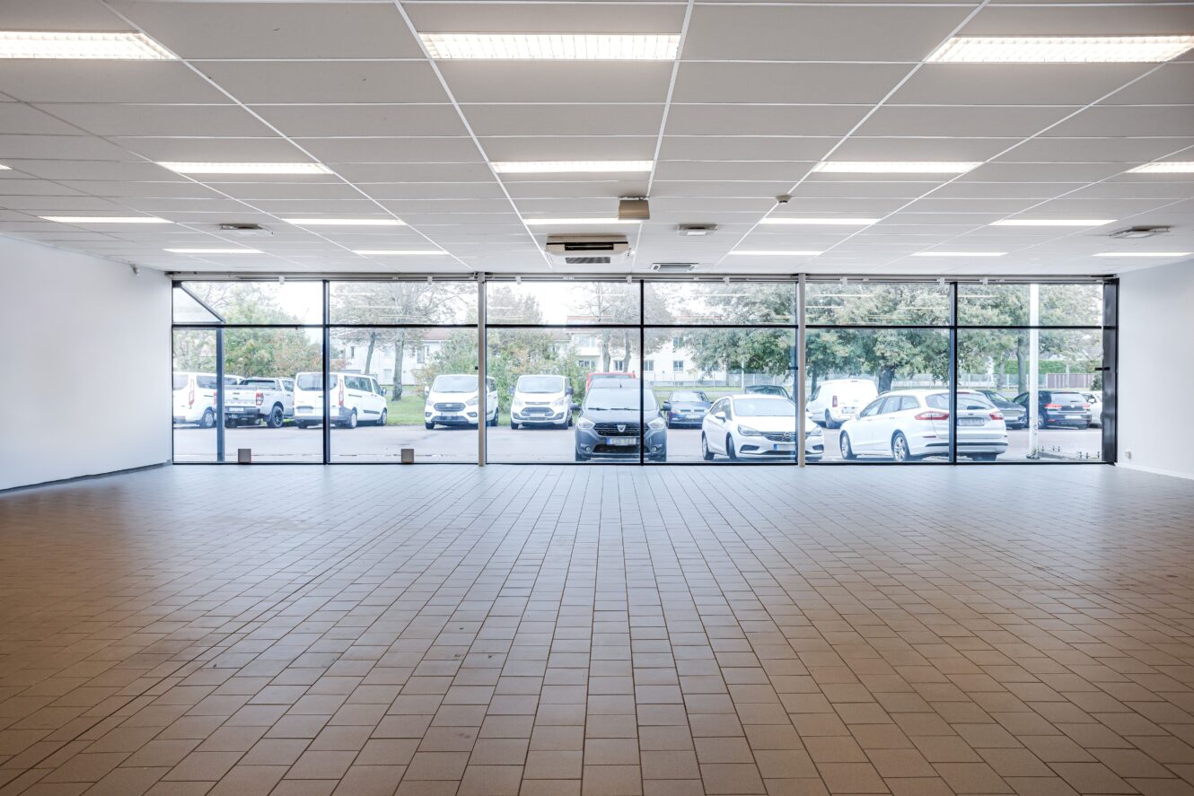 Tomt rum med klinkergolv och taklampor, mot stora fönster som visar flera parkerade bilar och träd utanför.