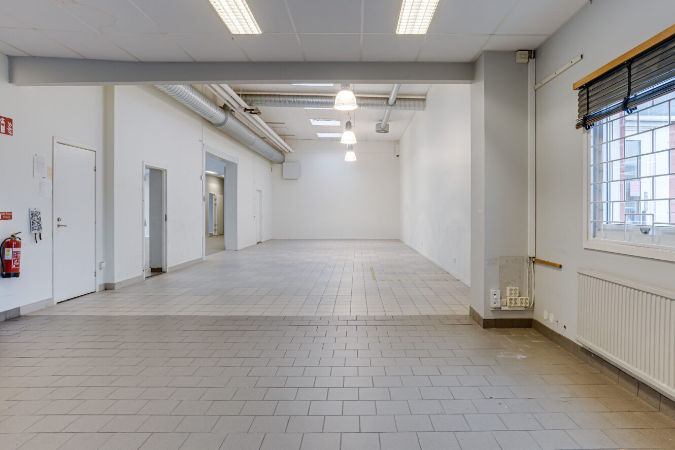 Tomt industriliknande rum med klinkergolv, vita väggar, takbelysning, synliga rör i taket, fönster med persienner och flera dörrar längs sidan.