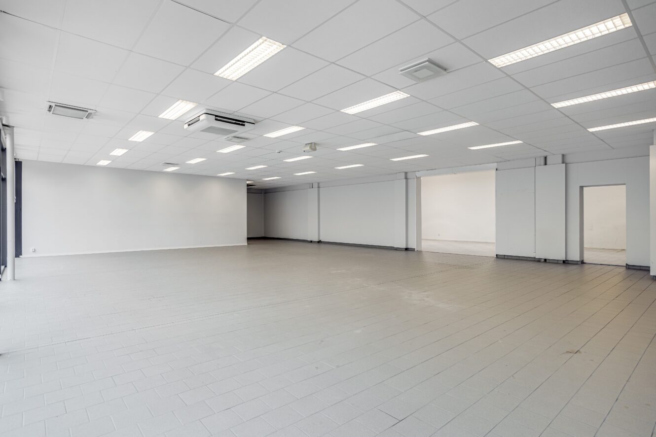Rymligt, tomt rum med klinkergolv, vita väggar och ett tak med lysrör och luftkonditioneringsenheter. Flera dörröppningar är synliga.