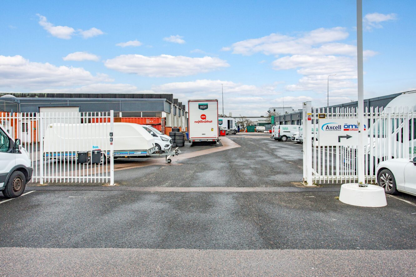 Gated entrance till ett industriområde med parkerade skåpbilar och lastbilar längs sidorna, omgiven av kommersiella byggnader under en delvis molnig himmel.