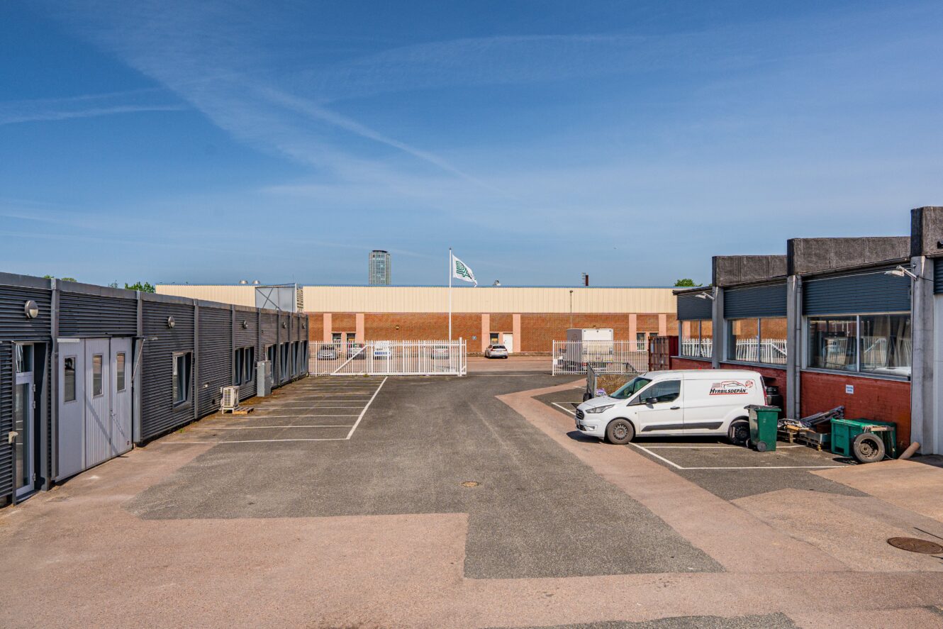 Industriområde med lagerbyggnader, en vit skåpbil parkerad till höger, tomma parkeringsplatser och en flagga på en stång i mitten under en klarblå himmel.