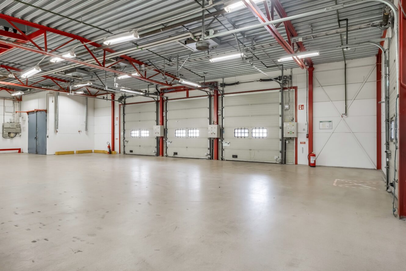 Inredning i ett tomt industriellt garage med fyra stora takskjutportar, synliga metallbalkar, stark belysning och ett polerat betonggolv.