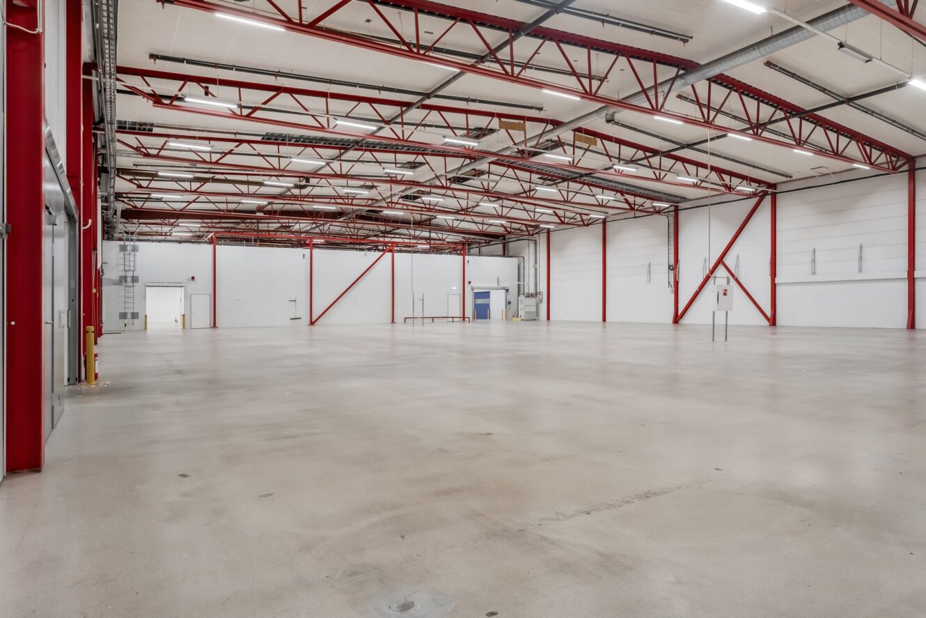 Rymlig, tom industrilagerlokal med högt i tak, röda stålbalkar, betonggolv och stora vita väggar.
