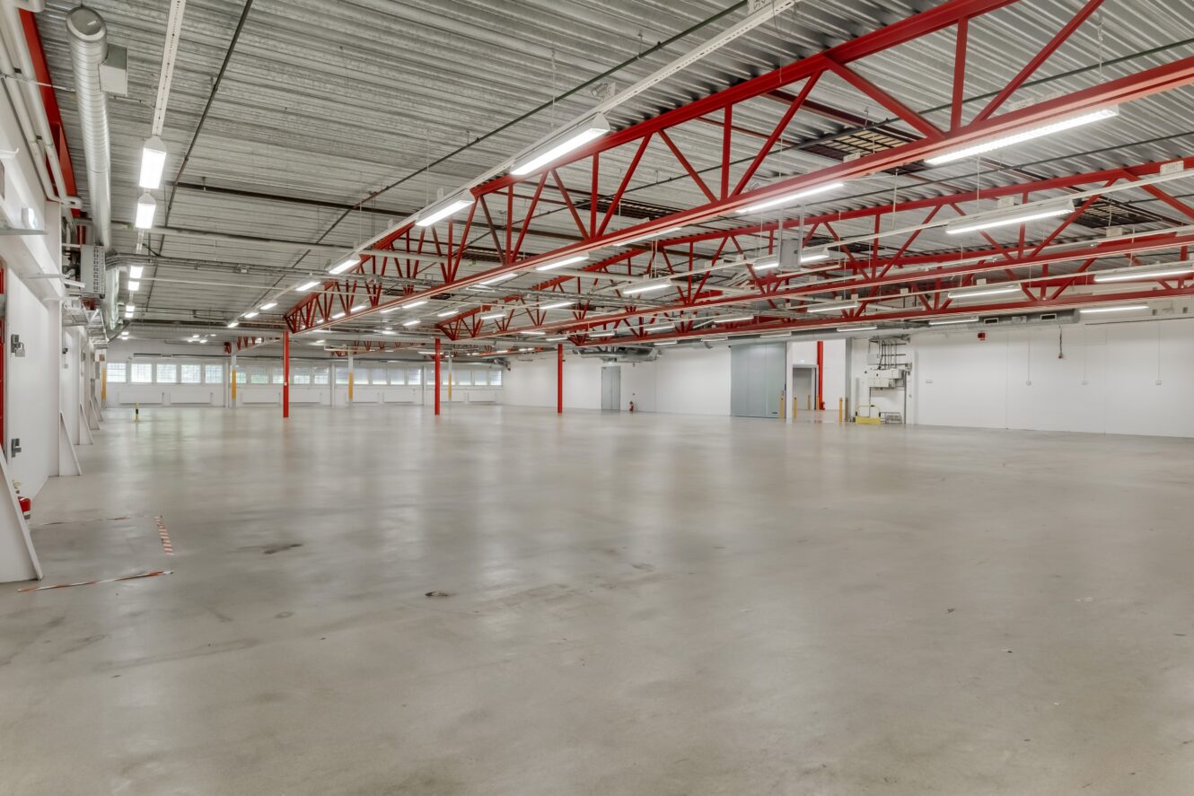 Rymlig, tom lagerlokal med betonggolv, vita väggar, röda stålbalkar och lysrör i taket. Stora fönster längs den bortre väggen.