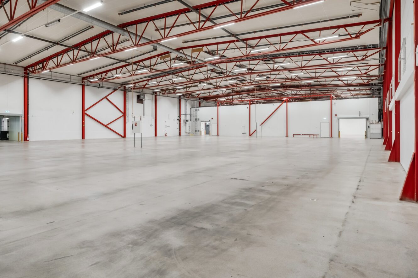 Stor tom lagerlokal med polerat betonggolv, vita väggar, röda stålbalkar och högt i tak med synliga rör och belysning.