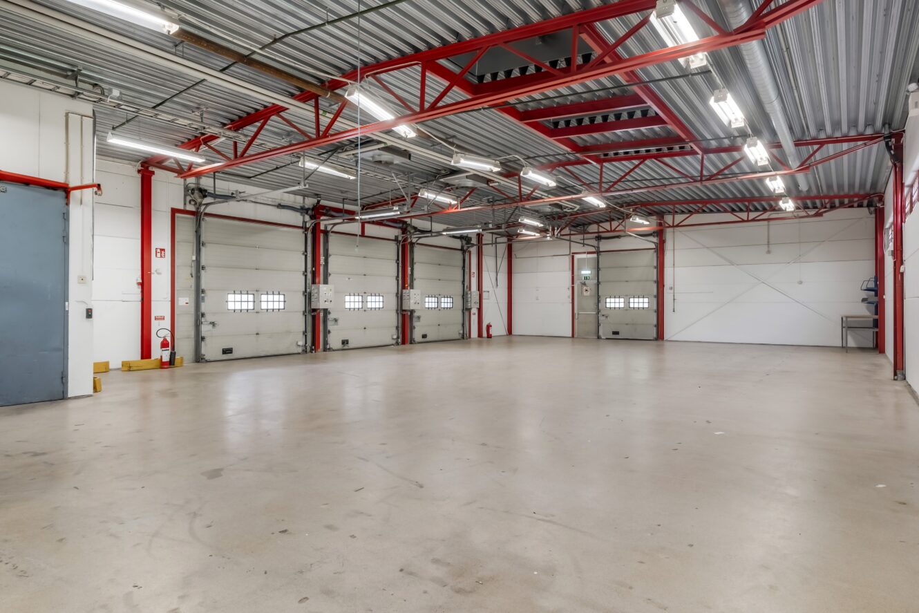 Rymligt, tomt industriellt garage med polerat betonggolv, röda stålbalkar, lysrörsbelysning och flera stora vita garageportar.