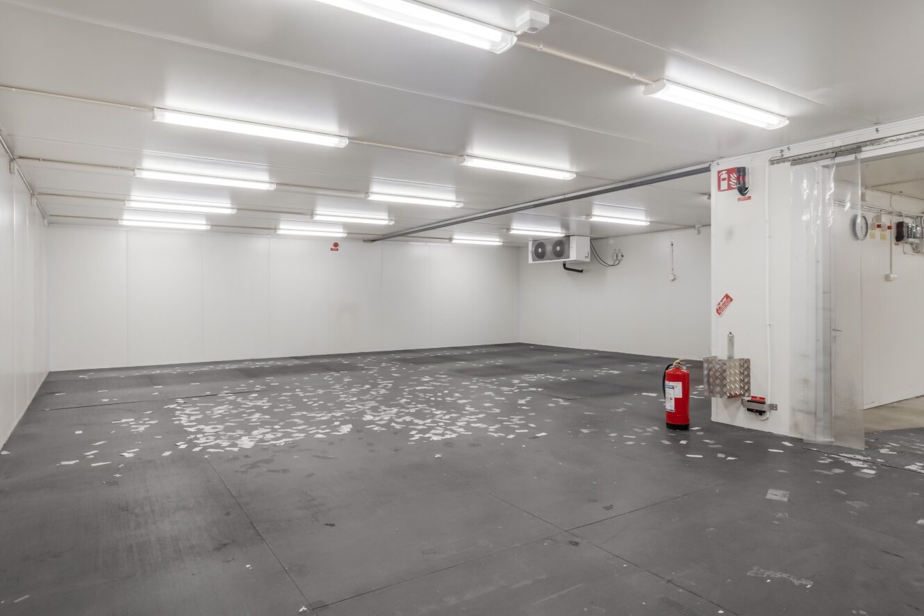 Tomt industrirum med svart golv, vita väggar och tak, lysrör i taket, skräp på golvet och en brandsläckare vid ett genomskinligt plastdraperi.