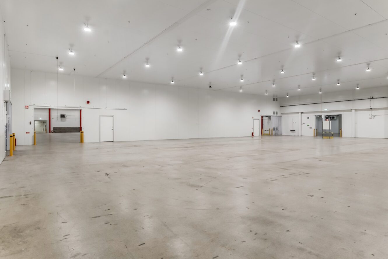 Stort, tomt industrilager med polerat betonggolv, vita väggar, taklampor och lastbryggor i bakgrunden.