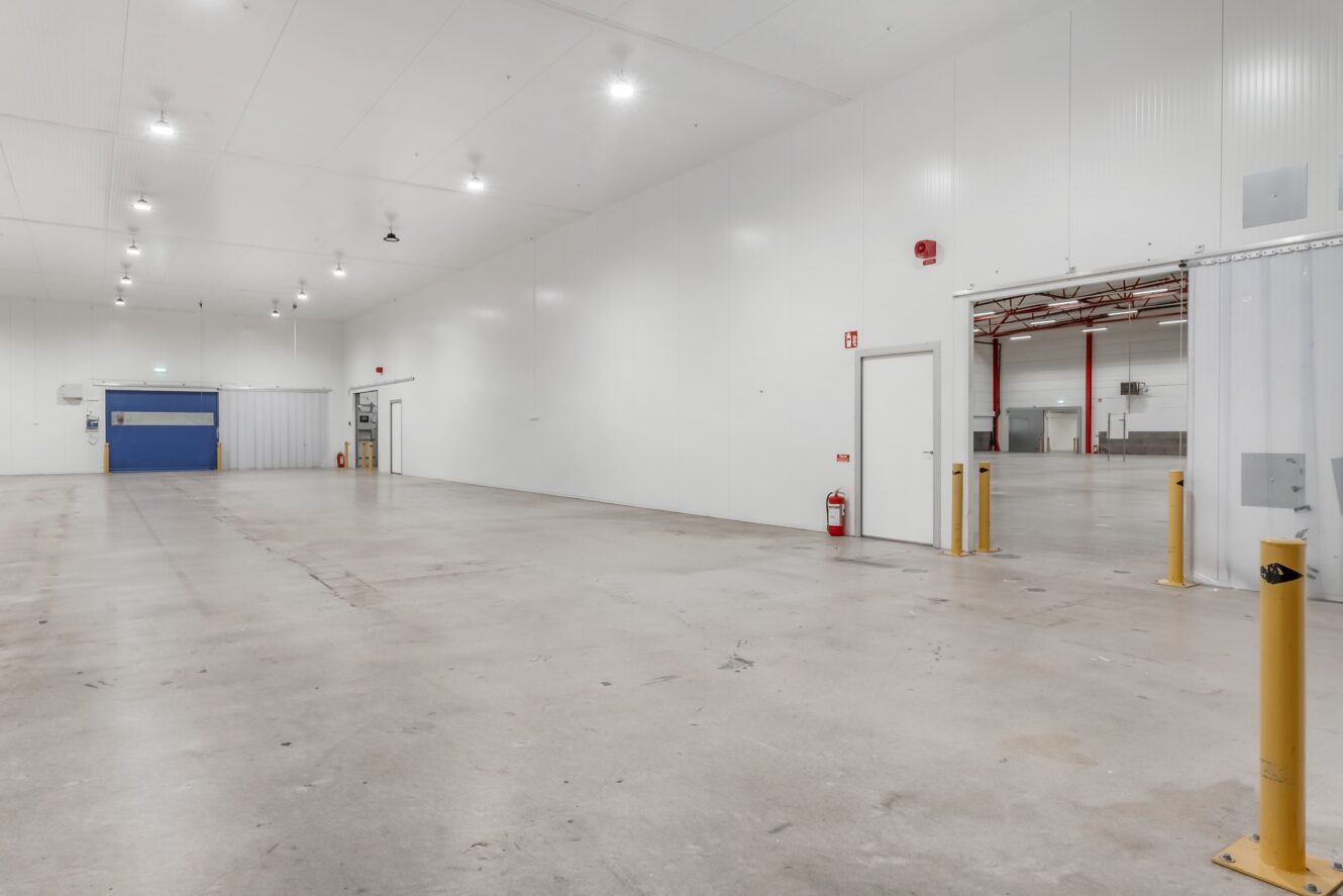 Stort tomt industrilager med vita väggar, betonggolv, takbelysning, en blå garageport och säkerhetsdetaljer som pollare och en brandsläckare.