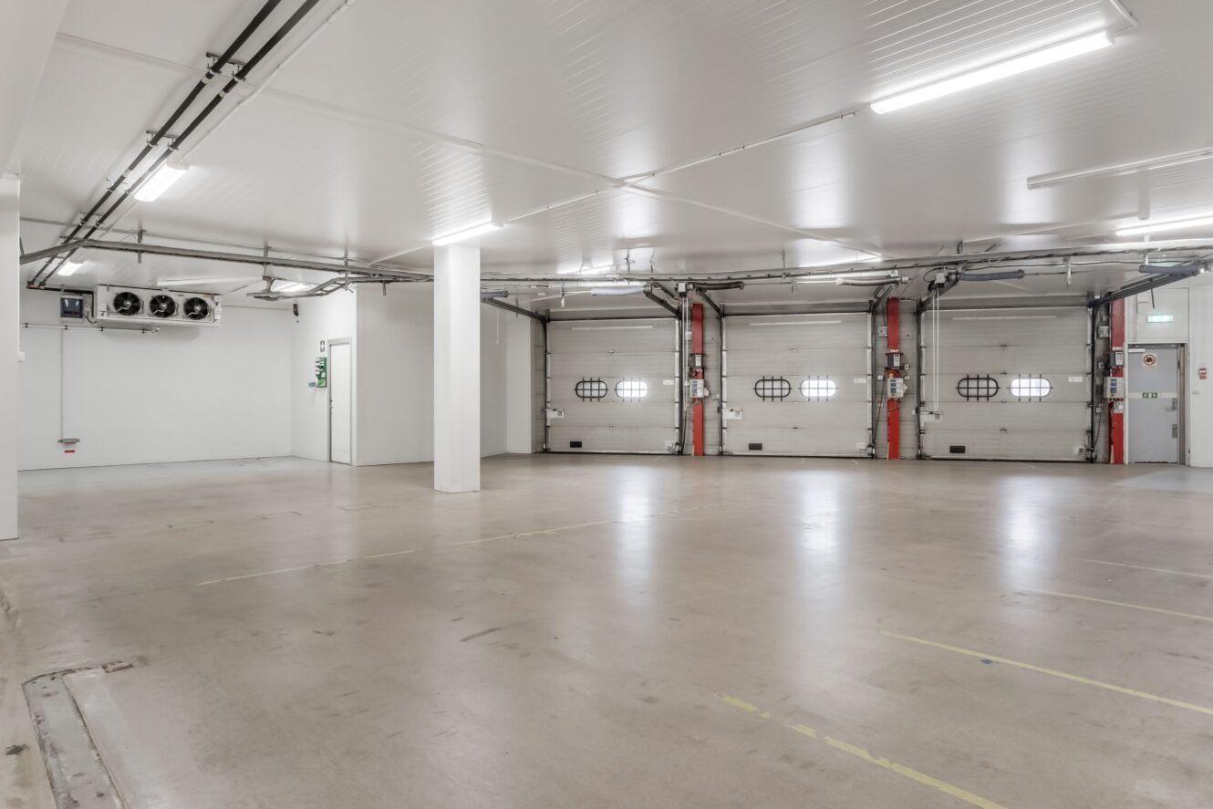 Ett tomt, rent industriellt lagerutrymme med vita väggar, ett slätt betonggolv och flera stängda garageportar längs ena sidan.
