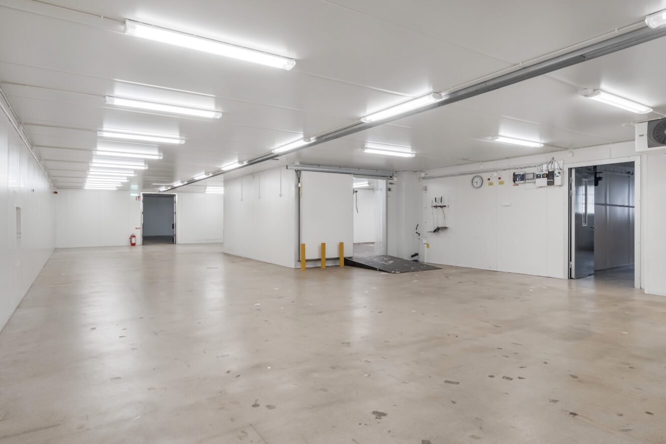 Rymlig, tom industrilokal med vita väggar, stark takbelysning, en ramp, verktygsutrustning och synliga dörrar som leder till andra utrymmen.