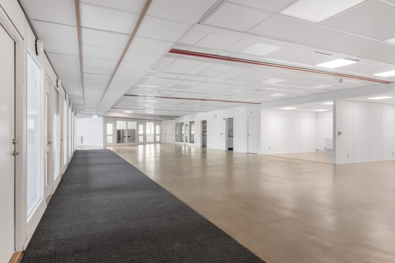 Rymlig, tom butikslokal med vita väggar, kaklat tak, stora fönster, beige golv och en del mörk heltäckningsmatta.