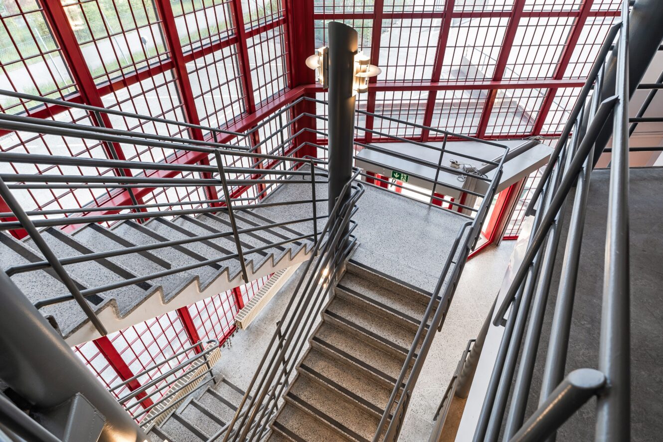 En inomhustrappa i flera plan med grå trappsteg och metallräcken, omgiven av stora glasfönster med röda ramar, sett uppifrån.