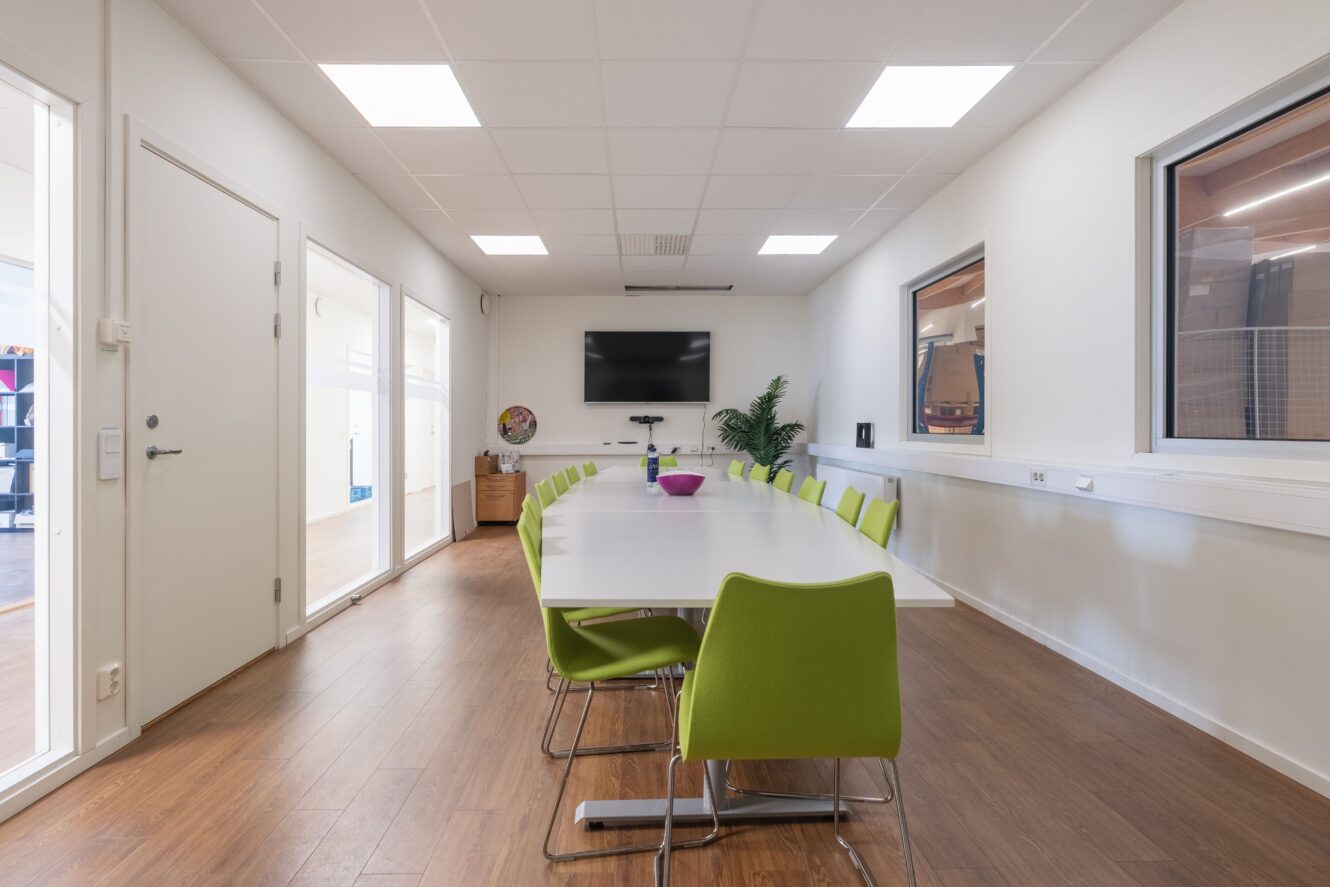 Modernt konferensrum med ett långt vitt bord, gröna stolar, en väggmonterad TV och stora fönster, trägolv och en krukväxt.