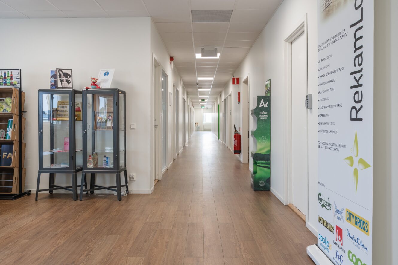 En ren kontorskorridor med trägolv, vita väggar, flera dörrar, en glasmonter till vänster och en hög reklambanner till höger.
