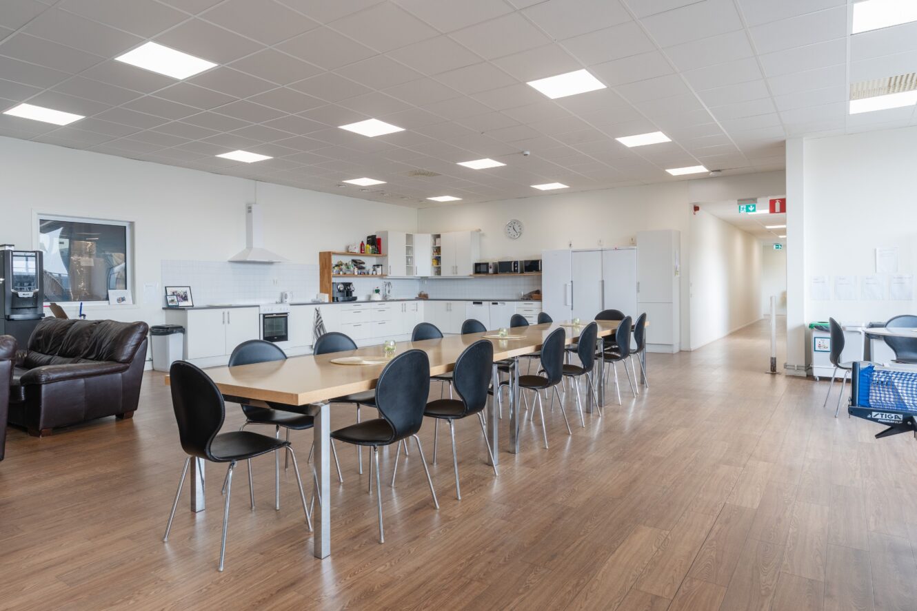 Ett rymligt, modernt kontorskök och matplats med ett långt bord, svarta stolar, vita skåp och trägolv.
