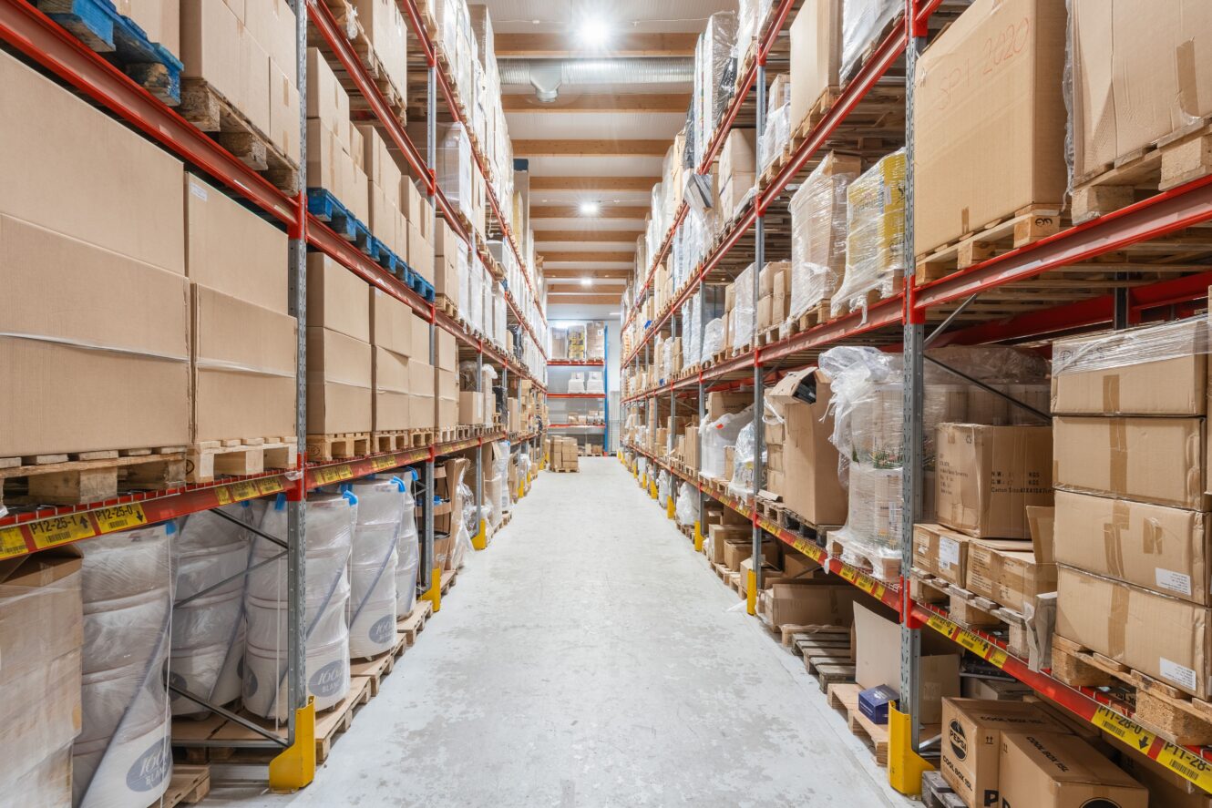 Bred lagergång med höga hyllor fulla av lådor och varor, upplysta av taklampor.