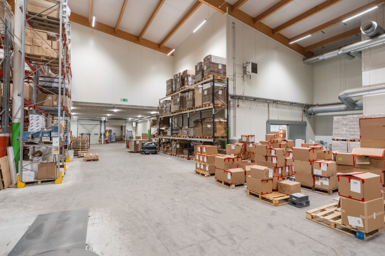 Rymlig lagerlokal med höga hyllor och pallar staplade med kartonger, högt i tak och organiserade förvaringsutrymmen.