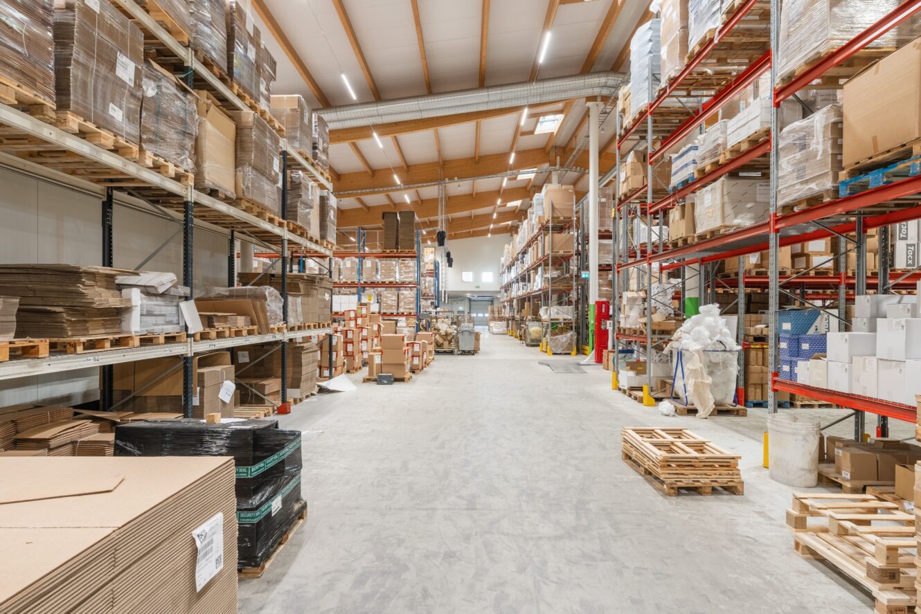 Bred lagergång med höga hyllor staplade med lådor, pallar och förpackningsmaterial under ett högt tak med takfönster.