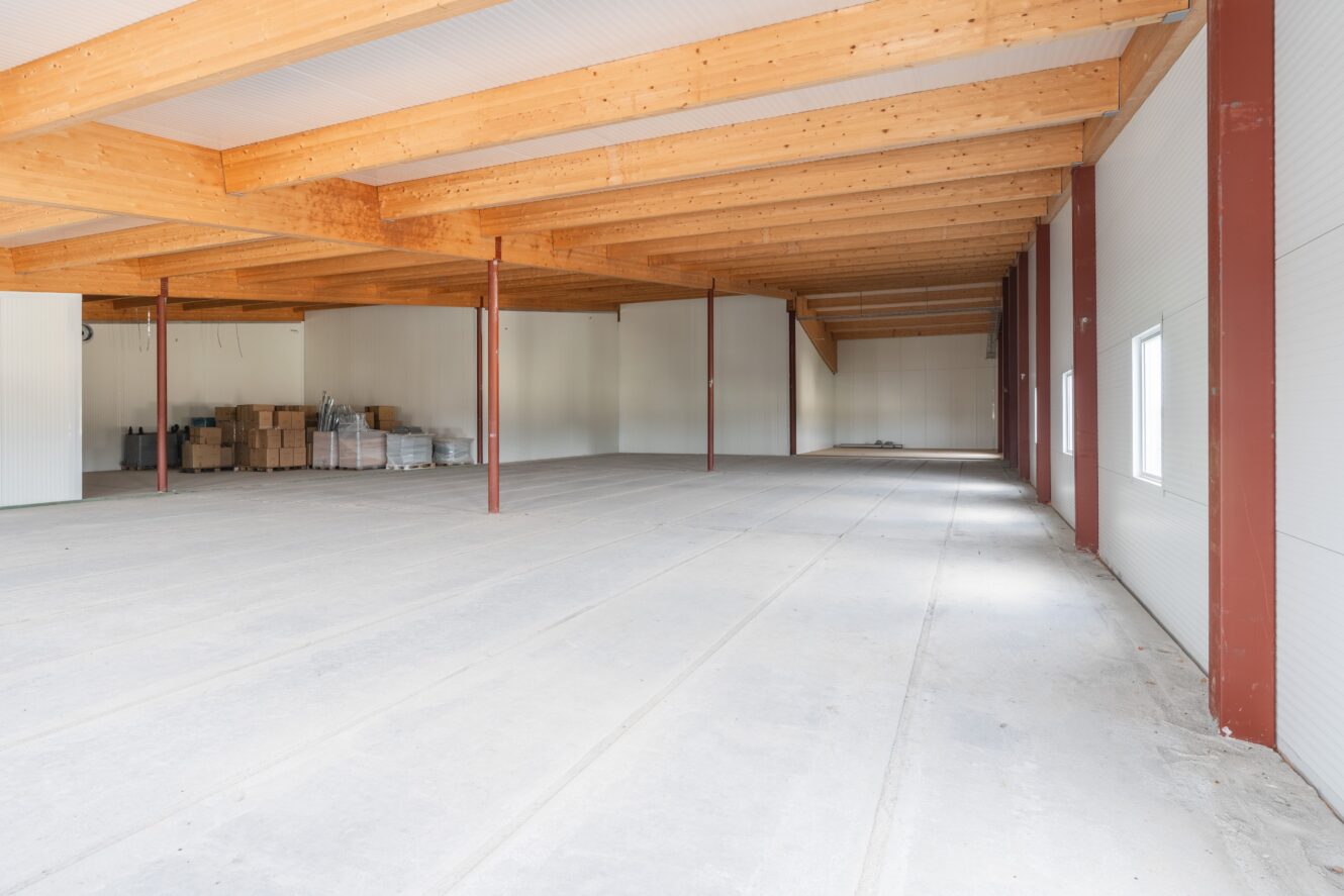 Rymlig, tom lagerlokal med synliga träbjälkar, vita väggar, stora fönster på ena sidan och staplade lådor i det bortre hörnet.