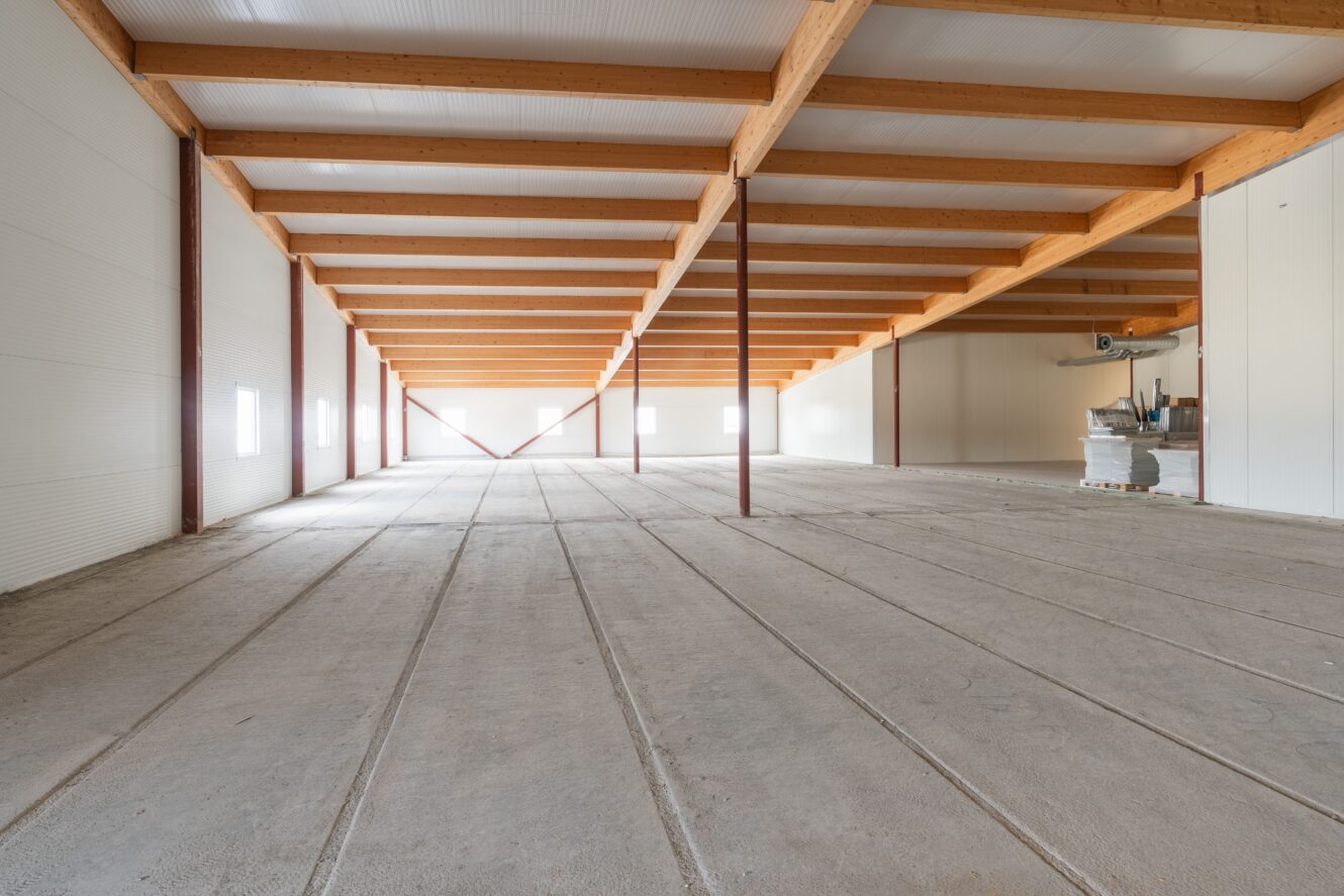 Stor, tom lagerlokal med träbjälkar, betonggolv, väggar med vit panel och staplade föremål i det bakre högra hörnet.