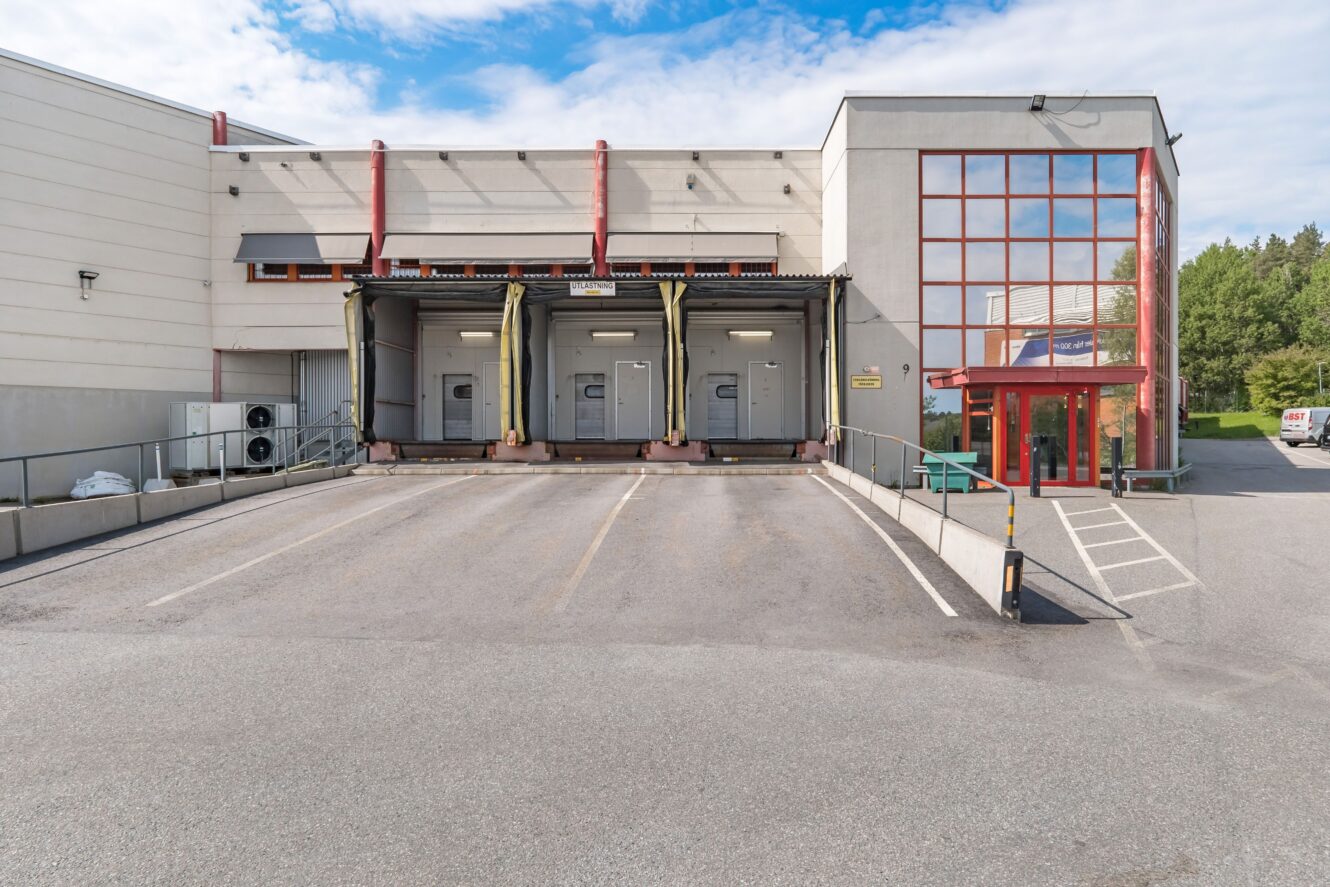 En lastkaj i ett lager med tre lastkajer, metallräcken och en glasentré på höger sida under en delvis molnig himmel.