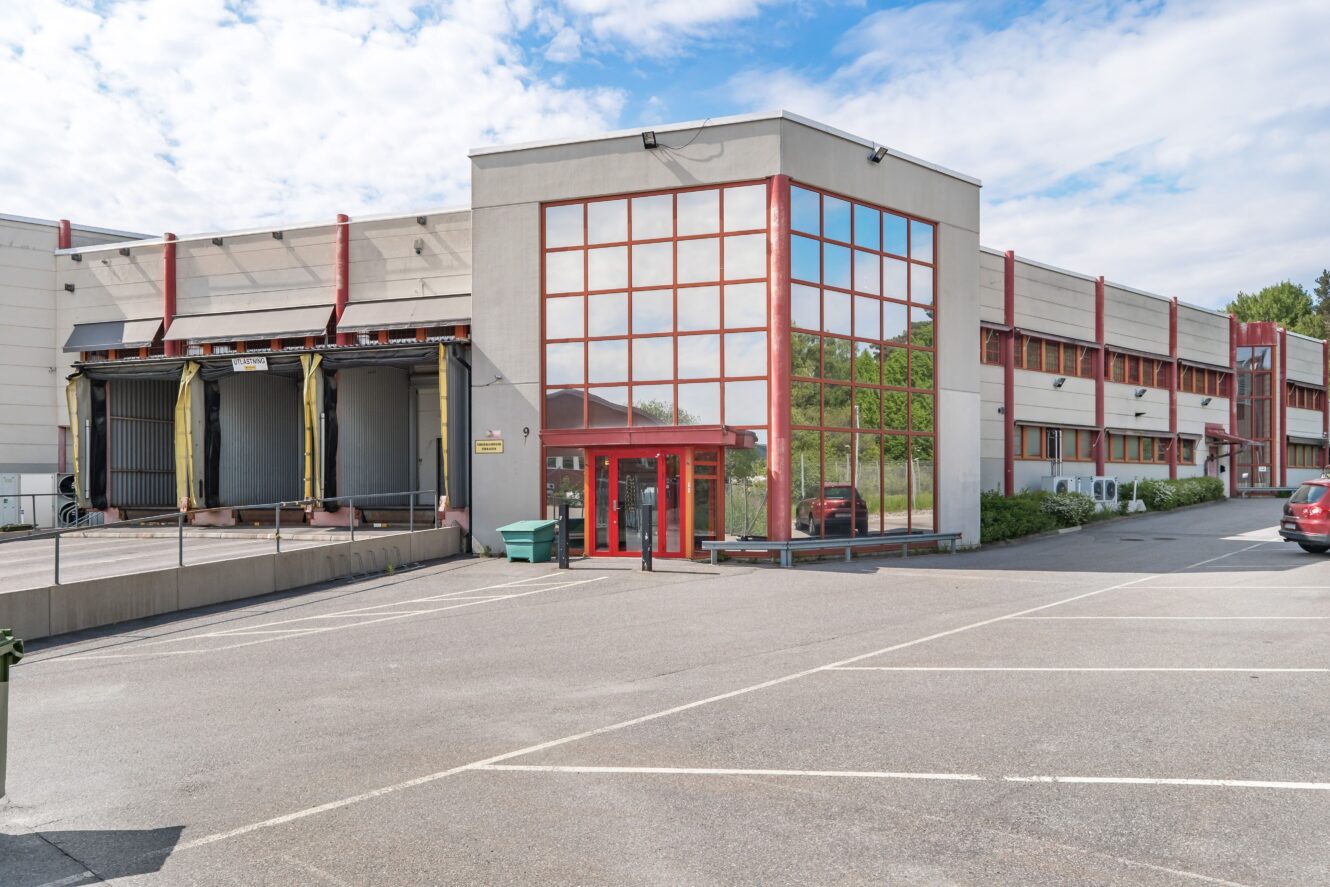 En stor industribyggnad med rödkarmade fönster, en lastkaj till vänster och en tom parkeringsplats framför under en delvis molnig himmel.