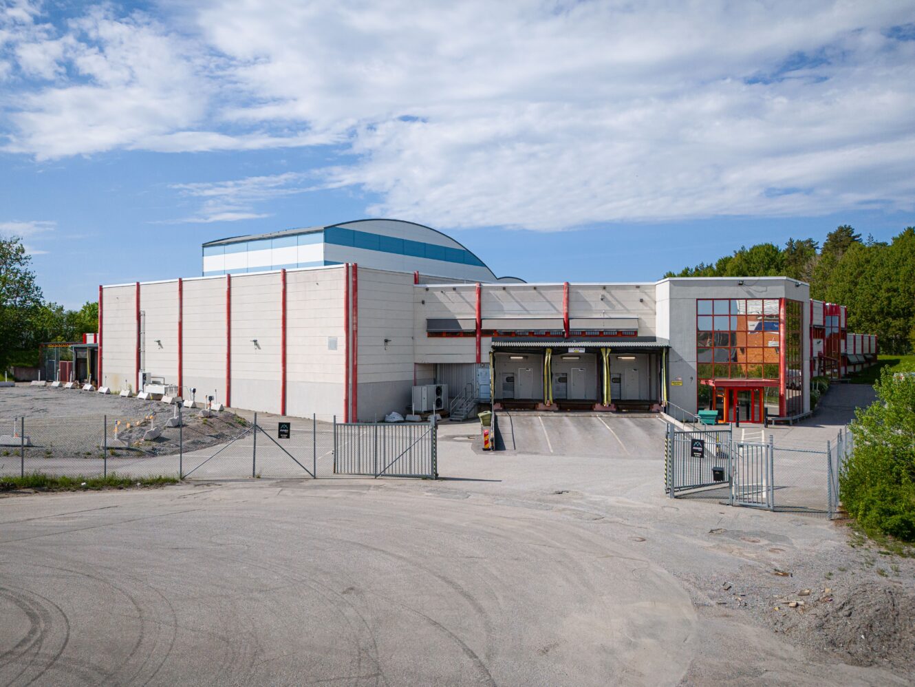 Ett stort, vitt industrilager med flera lastkajer, inhägnad tomt och omgivande grus och träd under en delvis molnig himmel.