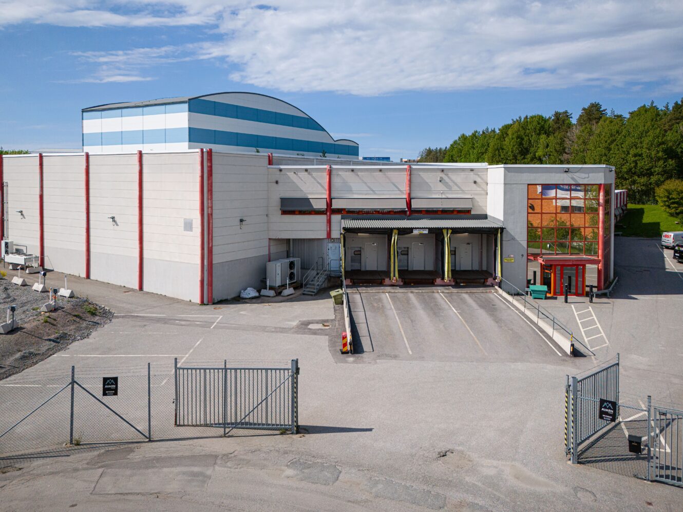 En inhägnad industribyggnad med fyra lastkajer, en glasad entré och en blandning av vita och blå ytterväggar, omgiven av träd och en tom gård.
