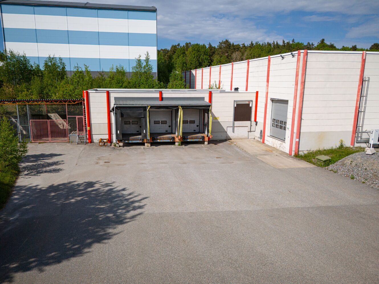 En lastkaj med tre lastbilsuppställningsplatser i anslutning till en ljus industribyggnad, omgiven av träd och ett inhägnat område till vänster.