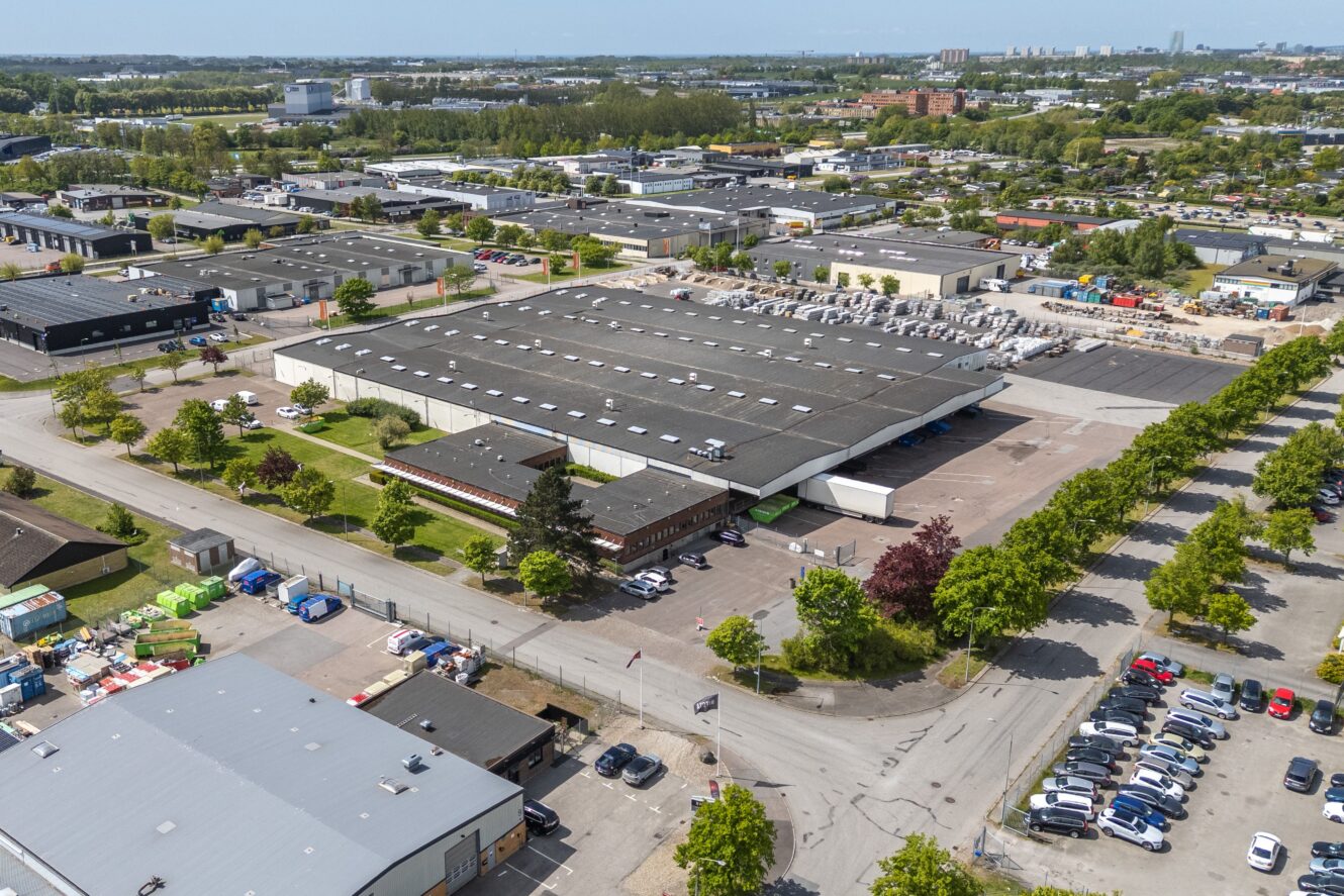 Flygfoto över ett stort industrilager med intilliggande parkeringsplatser, omgivet av träd och andra kommersiella byggnader i ett industriområde.