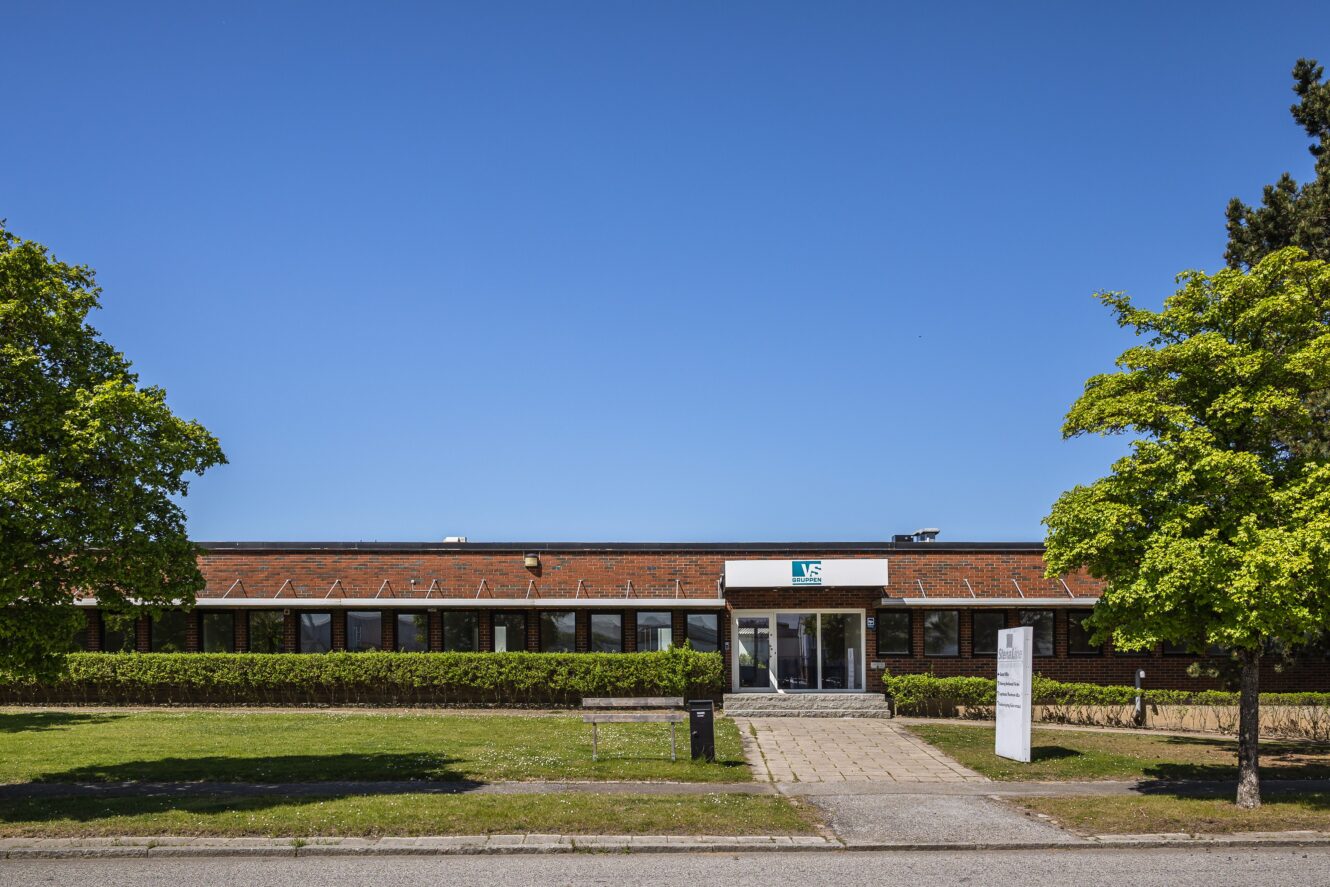 Kontorsbyggnad i en våning med rött tegeltak och glasad entré, omgiven av gröna träd och gräsmatta under klarblå himmel.