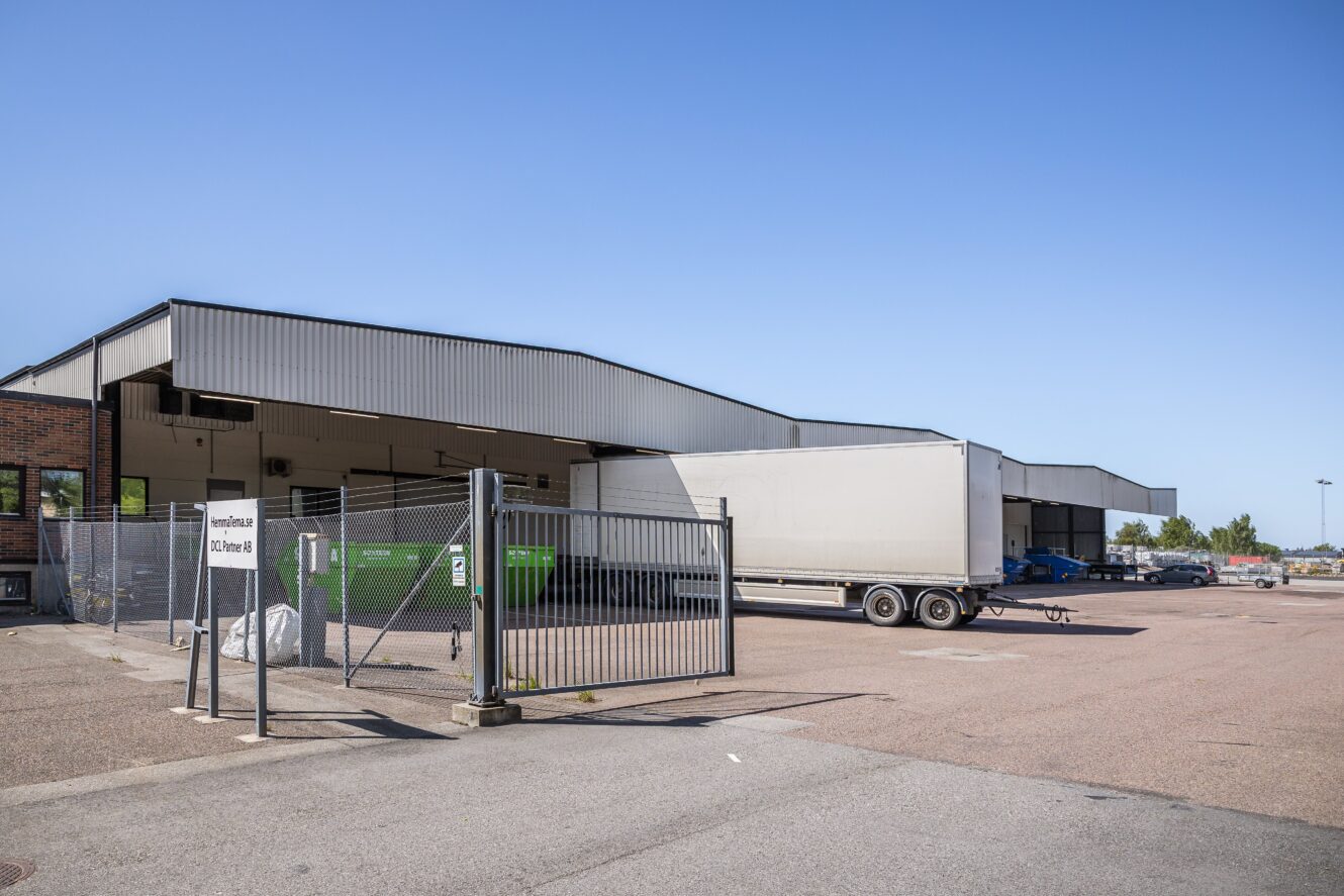 En inhägnad industriell lastkaj med flera kajer, släpvagnar parkerade vid kajerna och containrar för grönt avfall som syns en solig dag.